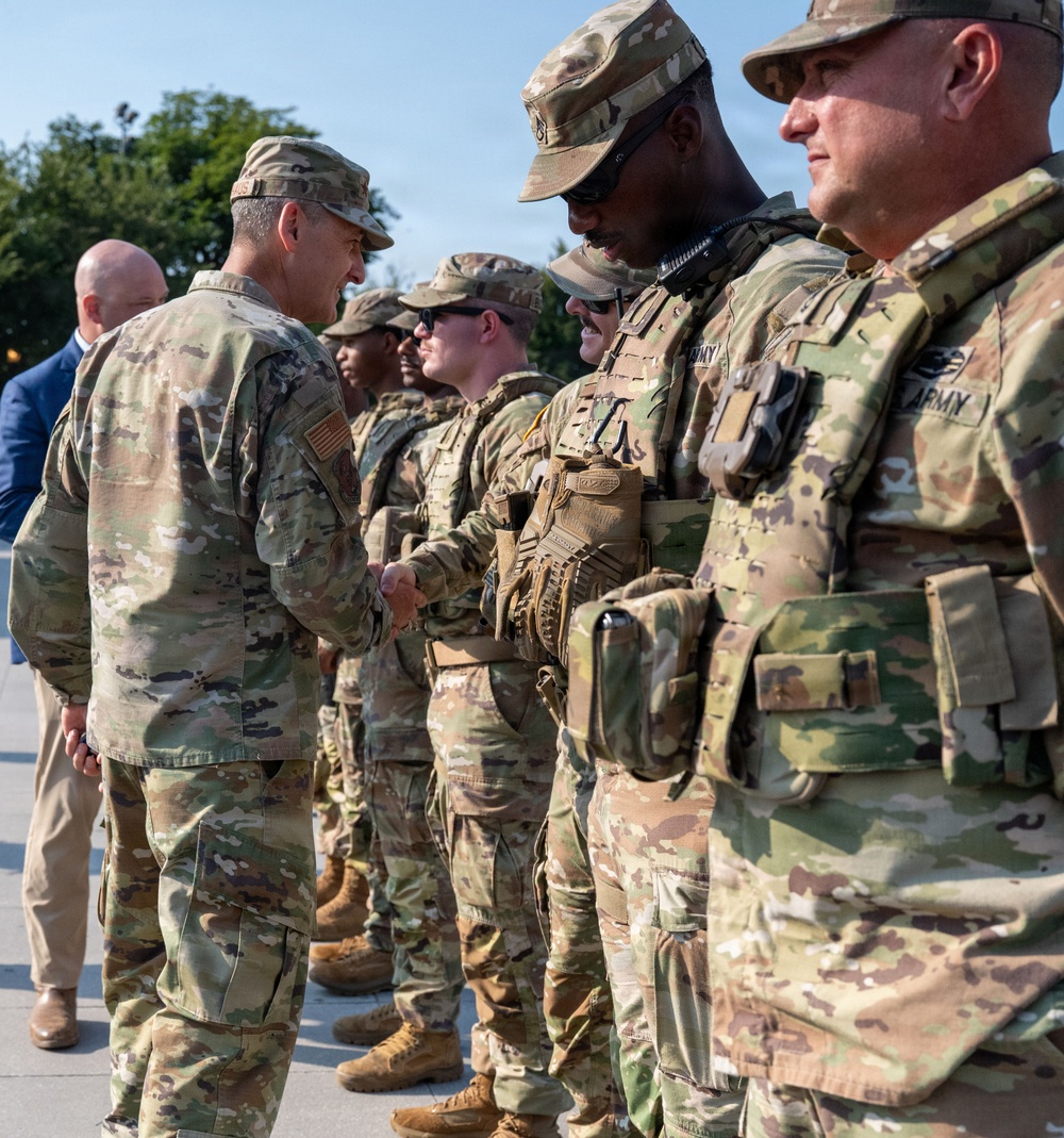 A senior military officer in U.S. Army Operational Camouflage Pattern (OCP) uniform is walking down a line of soldiers, shaking hands with them in what appears to be a formal greeting or recognition ceremony. The soldiers in the line are wearing full combat gear including body armor and tactical equipment. The setting appears to be an outdoor paved area with trees visible in the background.