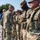 A senior military officer in U.S. Army Operational Camouflage Pattern (OCP) uniform is walking down a line of soldiers, shaking hands with them in what appears to be a formal greeting or recognition ceremony. The soldiers in the line are wearing full combat gear including body armor and tactical equipment. The setting appears to be an outdoor paved area with trees visible in the background.