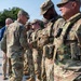 A senior military officer in U.S. Army Operational Camouflage Pattern (OCP) uniform is walking down a line of soldiers, shaking hands with them in what appears to be a formal greeting or recognition ceremony. The soldiers in the line are wearing full combat gear including body armor and tactical equipment. The setting appears to be an outdoor paved area with trees visible in the background.
