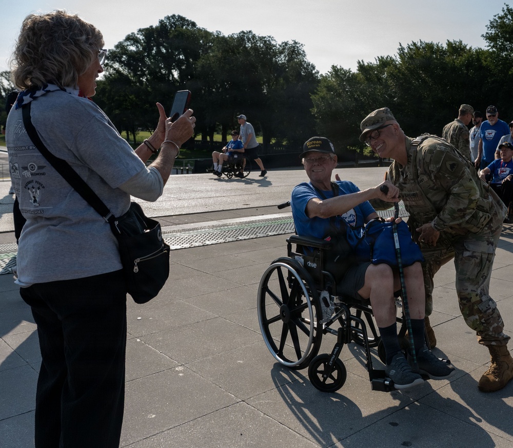 A Korean War veteran seated in a wheelchair poses for a photo with a U.S. Army soldier in multicam uniform, while a woman photographs them with a smartphone. The scene appears to take place at an outdoor memorial or public plaza, likely during an organized veterans' honor flight or tribute event. Additional participants in blue shirts and other veterans are visible in the background.