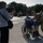 A Korean War veteran seated in a wheelchair poses for a photo with a U.S. Army soldier in multicam uniform, while a woman photographs them with a smartphone. The scene appears to take place at an outdoor memorial or public plaza, likely during an organized veterans' honor flight or tribute event. Additional participants in blue shirts and other veterans are visible in the background.