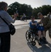 A Korean War veteran seated in a wheelchair poses for a photo with a U.S. Army soldier in multicam uniform, while a woman photographs them with a smartphone. The scene appears to take place at an outdoor memorial or public plaza, likely during an organized veterans' honor flight or tribute event. Additional participants in blue shirts and other veterans are visible in the background.