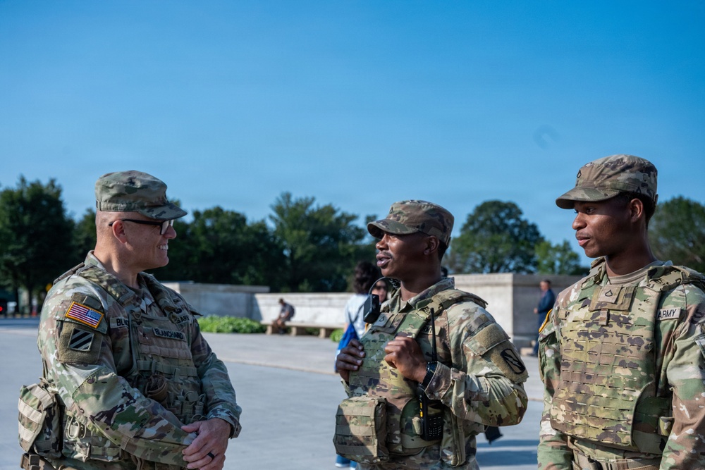 Three U.S. Army soldiers in Operational Camouflage Pattern (OCP) uniforms are engaged in conversation outdoors in what appears to be a public or ceremonial area. The soldier on the left is smiling and wearing name tapes and a 3rd Infantry Division patch. The setting appears to be a well-maintained outdoor plaza or memorial area with trees and clear blue sky in the background.