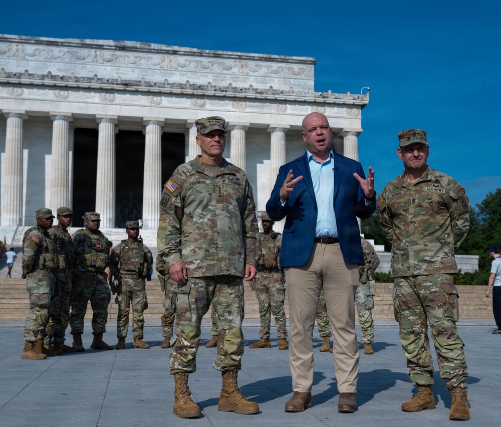 Three individuals stand in the foreground at the Lincoln Memorial in Washington, D.C. — two U.S. Army soldiers in OCP camouflage uniforms flanking a civilian in a blue blazer who appears to be speaking. In the background, additional uniformed soldiers are gathered near the memorial's iconic columns.