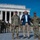 Three individuals stand in the foreground at the Lincoln Memorial in Washington, D.C. — two U.S. Army soldiers in OCP camouflage uniforms flanking a civilian in a blue blazer who appears to be speaking. In the background, additional uniformed soldiers are gathered near the memorial's iconic columns.