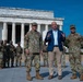Three individuals stand in the foreground at the Lincoln Memorial in Washington, D.C. — two U.S. Army soldiers in OCP camouflage uniforms flanking a civilian in a blue blazer who appears to be speaking. In the background, additional uniformed soldiers are gathered near the memorial's iconic columns.