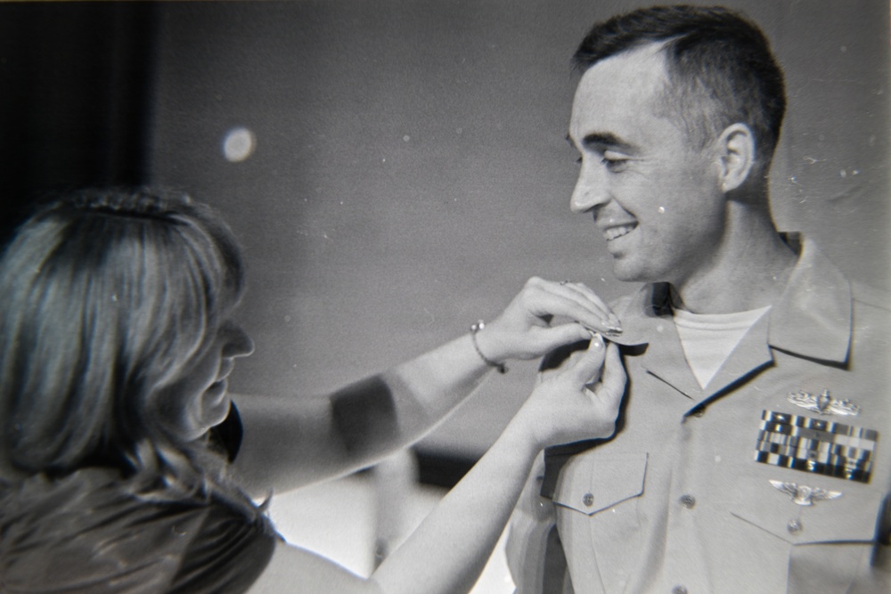 A black-and-white photograph showing a woman pinning a medal or insignia onto a smiling military officer in uniform. The officer is wearing a service uniform with multiple ribbon bars and what appears to be aviator wings, suggesting a promotion or award ceremony. The image appears to be from the mid-to-late 20th century based on photographic style and uniform details.