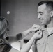 A black-and-white photograph showing a woman pinning a medal or insignia onto a smiling military officer in uniform. The officer is wearing a service uniform with multiple ribbon bars and what appears to be aviator wings, suggesting a promotion or award ceremony. The image appears to be from the mid-to-late 20th century based on photographic style and uniform details.