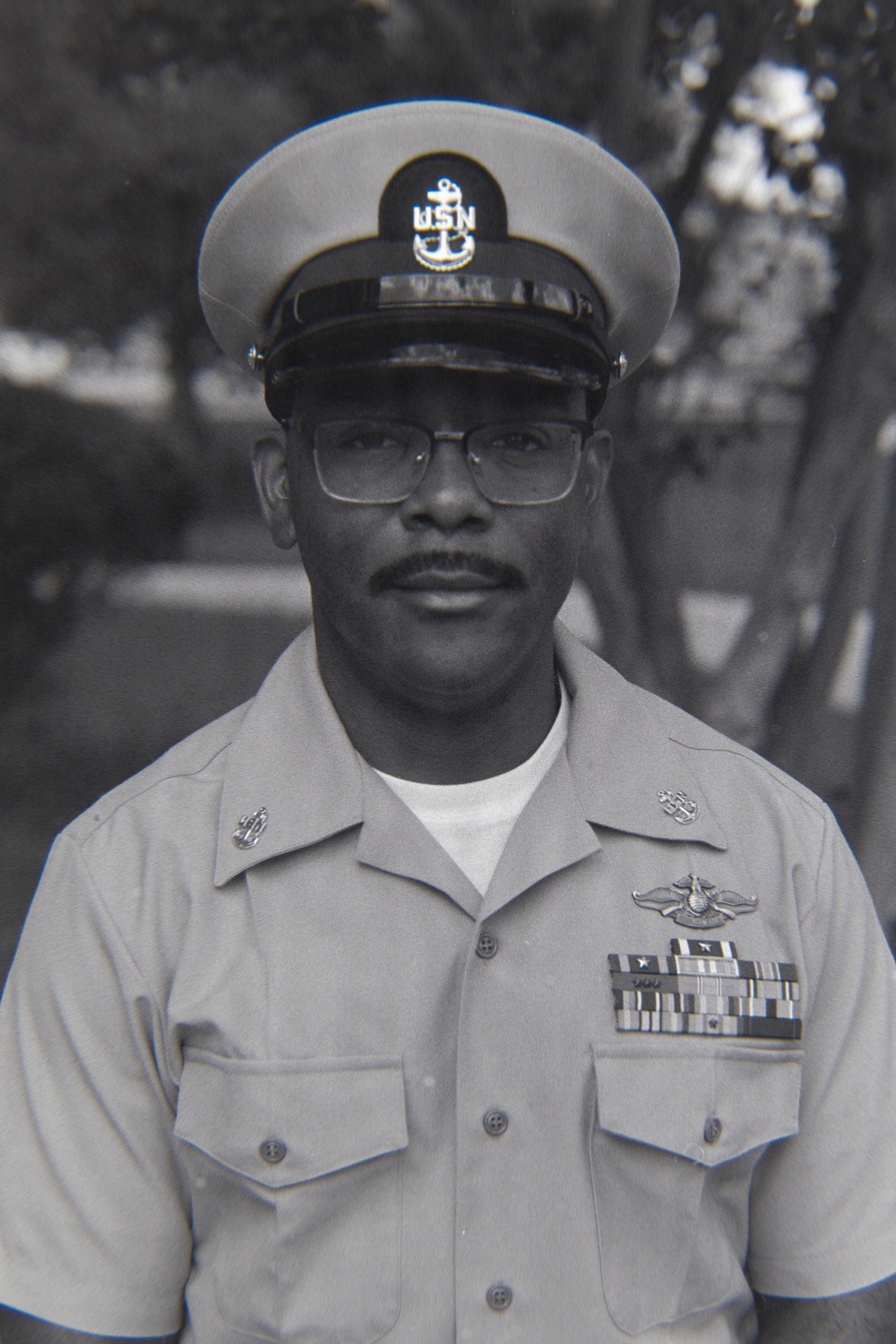 A black-and-white portrait photograph of a United States Navy Chief Petty Officer in dress uniform. The subject is wearing a combination cover (cap) with the USN anchor insignia, a short-sleeve khaki uniform with multiple ribbons, warfare devices, and collar insignia. The photo appears to be an official or semi-official military portrait taken outdoors.