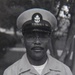 A black-and-white portrait photograph of a United States Navy Chief Petty Officer in dress uniform. The subject is wearing a combination cover (cap) with the USN anchor insignia, a short-sleeve khaki uniform with multiple ribbons, warfare devices, and collar insignia. The photo appears to be an official or semi-official military portrait taken outdoors.
