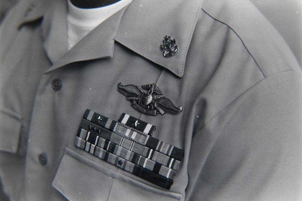 A close-up photograph of a U.S. Navy uniform shirt in gray/khaki color displaying multiple military insignia and ribbons. The uniform features a Fleet Marine Force (FMF) badge, a collar device, and a ribbon rack with multiple service ribbons and device attachments. The image appears to be a black-and-white or desaturated archival photograph.