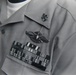 A close-up photograph of a U.S. Navy uniform shirt in gray/khaki color displaying multiple military insignia and ribbons. The uniform features a Fleet Marine Force (FMF) badge, a collar device, and a ribbon rack with multiple service ribbons and device attachments. The image appears to be a black-and-white or desaturated archival photograph.