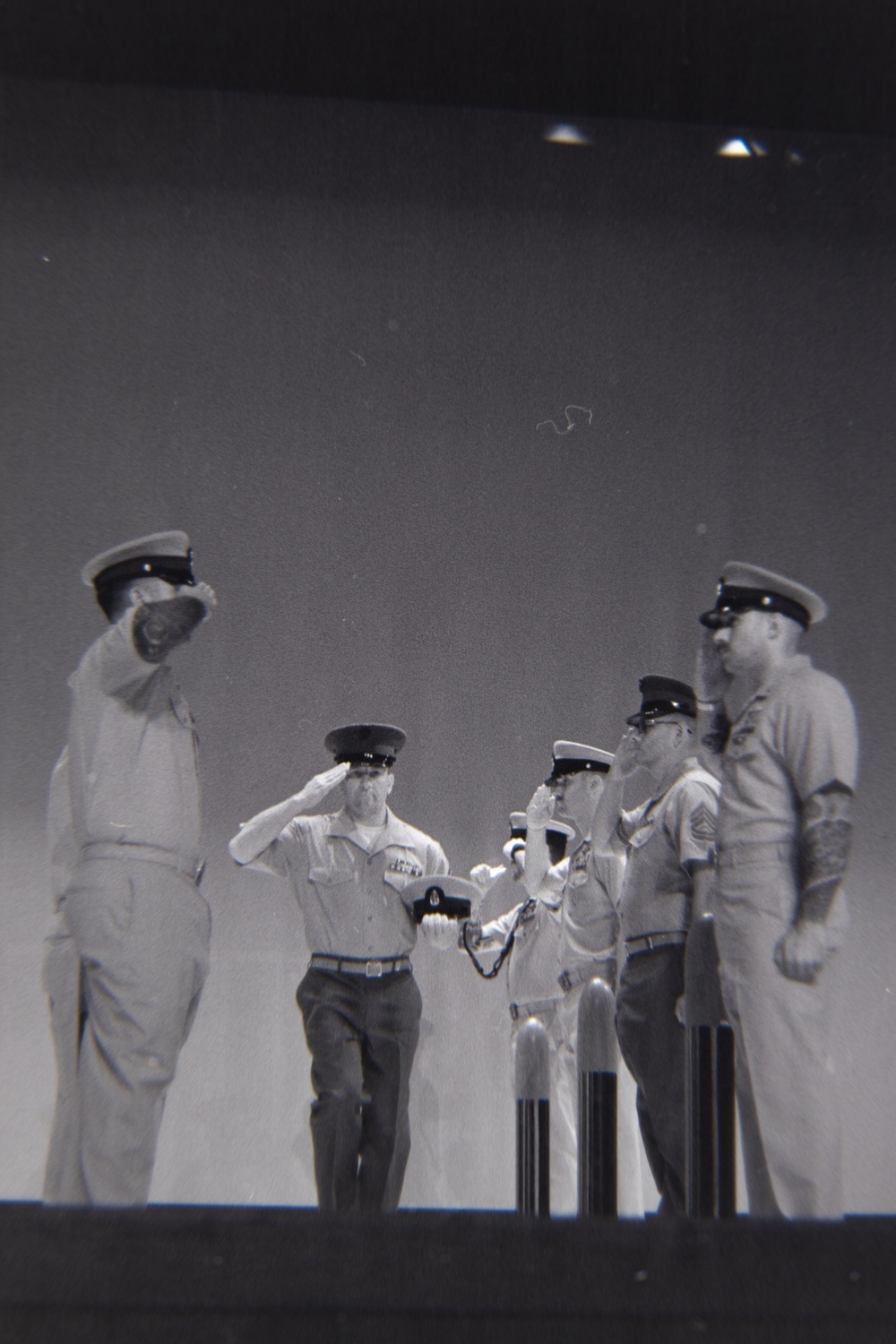 A black-and-white archival photograph, appearing to be a film negative or slide, depicting a group of military personnel in uniform on what appears to be a raised platform or stage. Several individuals are in saluting positions, suggesting a formal military ceremony or official event. The image shows characteristics of mid-20th century military photography.