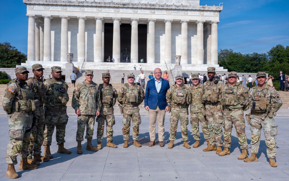 A group of approximately 12 military personnel in U.S. Army camouflage uniforms pose for a group photo with one civilian man in a blue blazer and khaki pants. The group is standing in front of the Lincoln Memorial in Washington, D.C., on a clear, sunny day. The iconic white marble columns and steps of the memorial are clearly visible in the background.