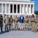 A group of approximately 12 military personnel in U.S. Army camouflage uniforms pose for a group photo with one civilian man in a blue blazer and khaki pants. The group is standing in front of the Lincoln Memorial in Washington, D.C., on a clear, sunny day. The iconic white marble columns and steps of the memorial are clearly visible in the background.
