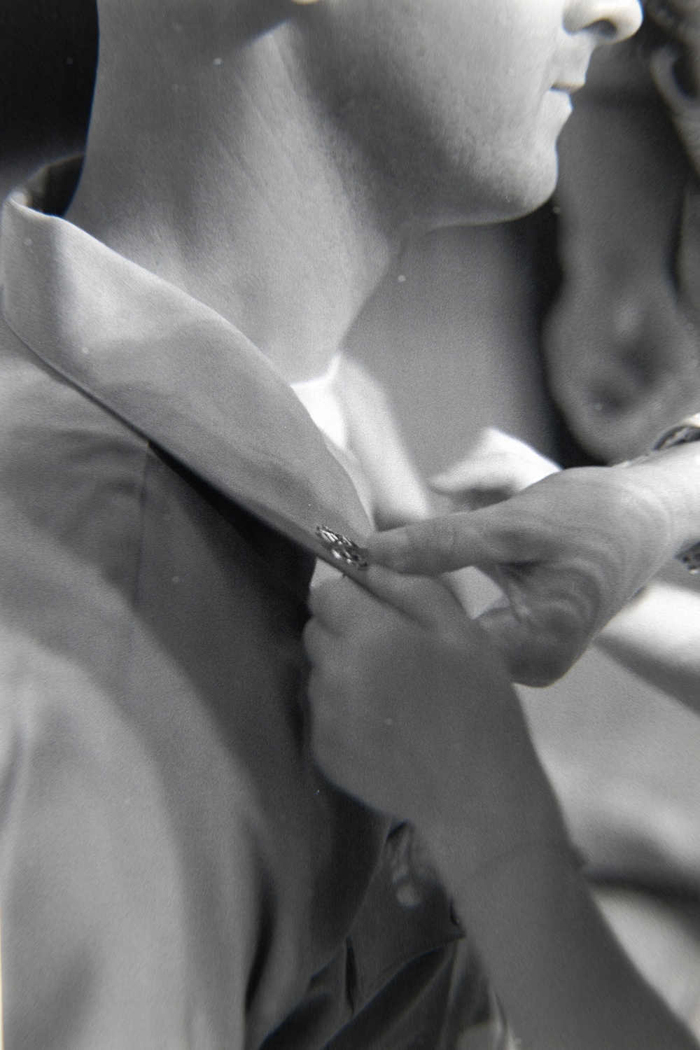 A black and white close-up photograph showing a person fastening or adjusting a cufflink on a dress shirt sleeve. A second person's hand is visible performing the action, suggesting assistance being given, possibly in a formal dressing context. The image has an intimate, candid quality consistent with event or wedding photography.