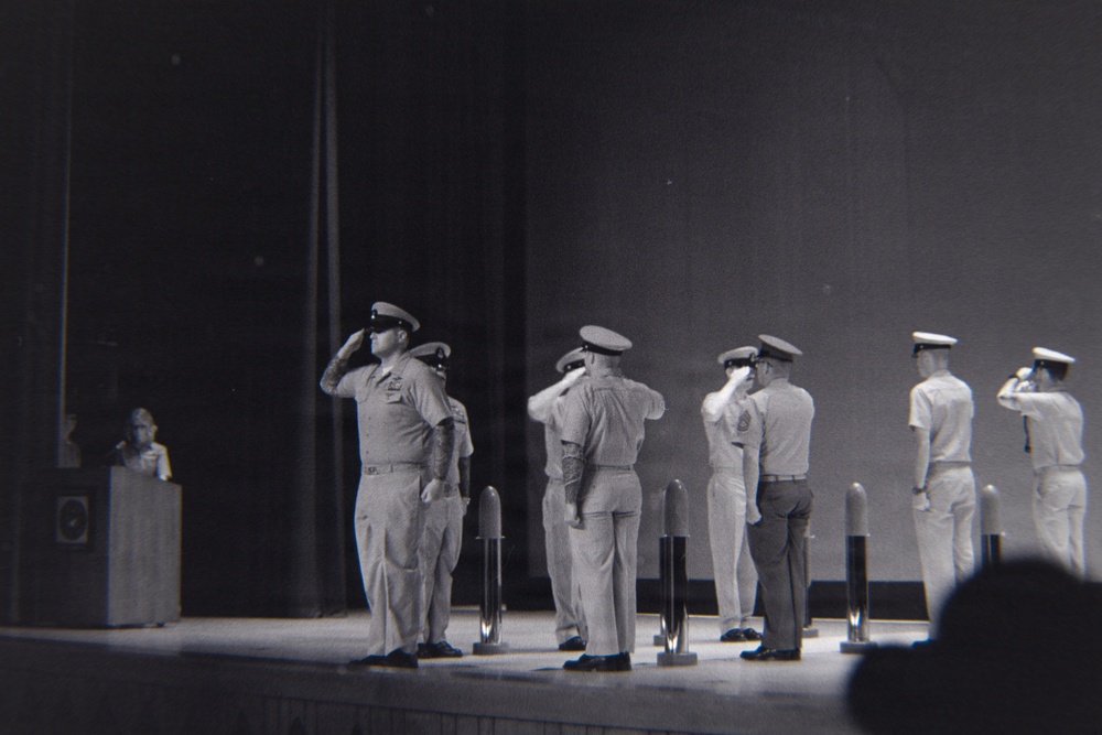 A black-and-white photograph depicting a formal military ceremony on a stage or platform, where multiple uniformed personnel are rendering salutes simultaneously. The individuals are wearing military dress uniforms with peaked caps, suggesting a structured official event such as a change of command, awards ceremony, or retirement ceremony. A figure at a podium is visible in the background left, indicating a formal program is in progress.