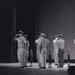 A black-and-white photograph depicting a formal military ceremony on a stage or platform, where multiple uniformed personnel are rendering salutes simultaneously. The individuals are wearing military dress uniforms with peaked caps, suggesting a structured official event such as a change of command, awards ceremony, or retirement ceremony. A figure at a podium is visible in the background left, indicating a formal program is in progress.