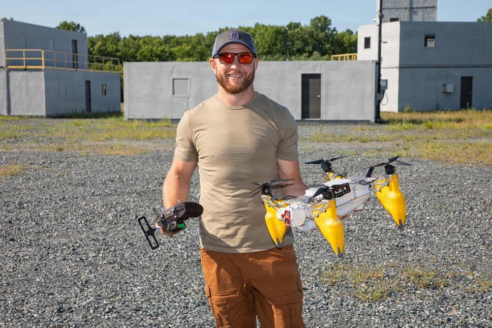 A bearded man wearing a tan t-shirt, brown cargo pants, red-tinted sunglasses, and a gray cap stands outdoors on a gravel surface holding two pieces of drone/UAV equipment. In his right hand he holds a white and yellow multi-rotor drone with pontoon-style floats, and in his left hand he holds what appears to be a remote controller or sensor module. The background features concrete block structures, a gravel lot, and green trees under a clear blue sky.