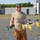 xTechAutonomy Phase II Live Demonstrations A bearded man wearing a tan t-shirt, brown cargo pants, red-tinted sunglasses, and a gray cap stands outdoors on a gravel surface holding two pieces of drone/UAV equipment. In his right hand he holds a white and yellow multi-rotor drone with pontoon-style floats, and in his left hand he holds what appears to be a remote controller or sensor module. The background features concrete block structures, a gravel lot, and green trees under a clear blue sky.