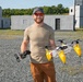 A bearded man wearing a tan t-shirt, brown cargo pants, red-tinted sunglasses, and a gray cap stands outdoors on a gravel surface holding two pieces of drone/UAV equipment. In his right hand he holds a white and yellow multi-rotor drone with pontoon-style floats, and in his left hand he holds what appears to be a remote controller or sensor module. The background features concrete block structures, a gravel lot, and green trees under a clear blue sky.