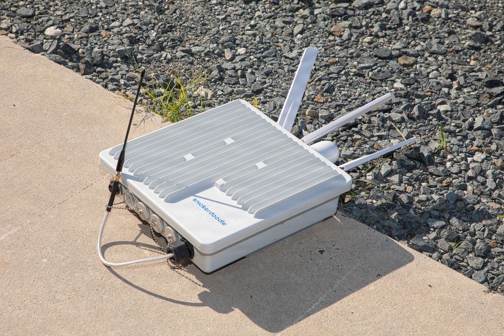 The image shows a white outdoor networking or wireless communication device branded 'snickerdoodle' placed on a concrete surface next to a gravel area. The device features multiple antennas extending from its body, along with cables and connectors attached to its side. It appears to be a ruggedized, weatherproof enclosure designed for outdoor deployment.