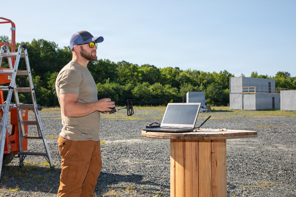 A bearded man wearing a baseball cap, sunglasses, olive t-shirt, and brown cargo pants stands outdoors on a gravel surface holding what appears to be a remote control or handheld device. He is positioned next to a wooden cable spool repurposed as a standing table, on which a laptop computer is open. The background shows a treeline, concrete block structures, an orange scissor lift, and a metal stepladder.