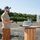 xTechAutonomy Phase II Live Demonstrations A bearded man wearing a baseball cap, sunglasses, olive t-shirt, and brown cargo pants stands outdoors on a gravel surface holding what appears to be a remote control or handheld device. He is positioned next to a wooden cable spool repurposed as a standing table, on which a laptop computer is open. The background shows a treeline, concrete block structures, an orange scissor lift, and a metal stepladder.