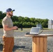 A bearded man wearing a baseball cap, sunglasses, olive t-shirt, and brown cargo pants stands outdoors on a gravel surface holding what appears to be a remote control or handheld device. He is positioned next to a wooden cable spool repurposed as a standing table, on which a laptop computer is open. The background shows a treeline, concrete block structures, an orange scissor lift, and a metal stepladder.