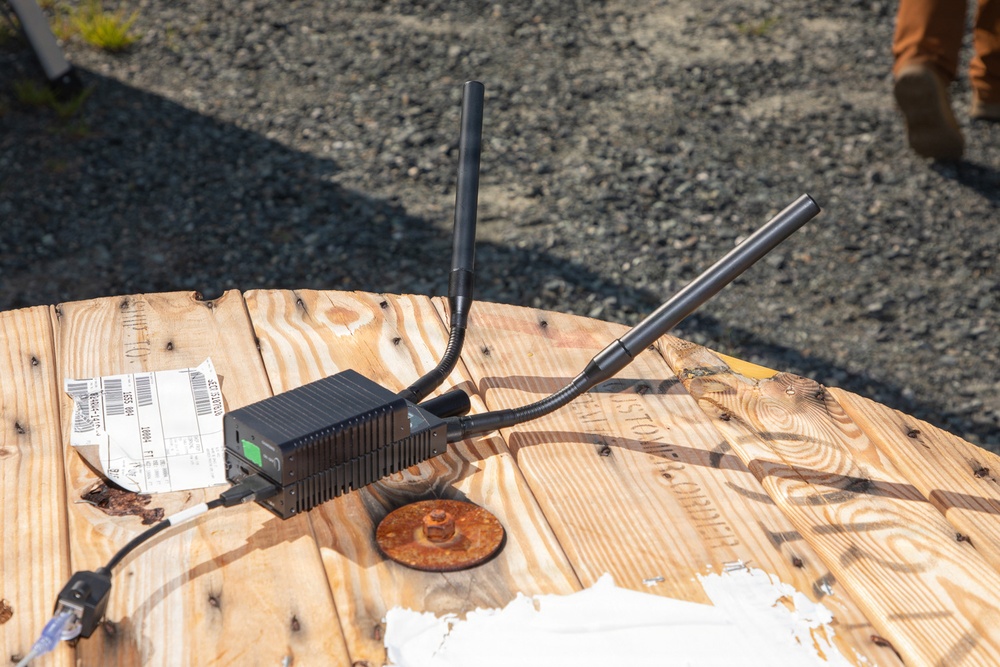A black wireless networking device with multiple external antennas is placed on top of a large wooden cable spool reel being used as an improvised table. The device appears to be a router or wireless access point deployed in an outdoor field setting, likely a construction or work site. A shipping label, ethernet cable, and miscellaneous papers are also visible on the wooden surface.