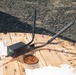 A black wireless networking device with multiple external antennas is placed on top of a large wooden cable spool reel being used as an improvised table. The device appears to be a router or wireless access point deployed in an outdoor field setting, likely a construction or work site. A shipping label, ethernet cable, and miscellaneous papers are also visible on the wooden surface.