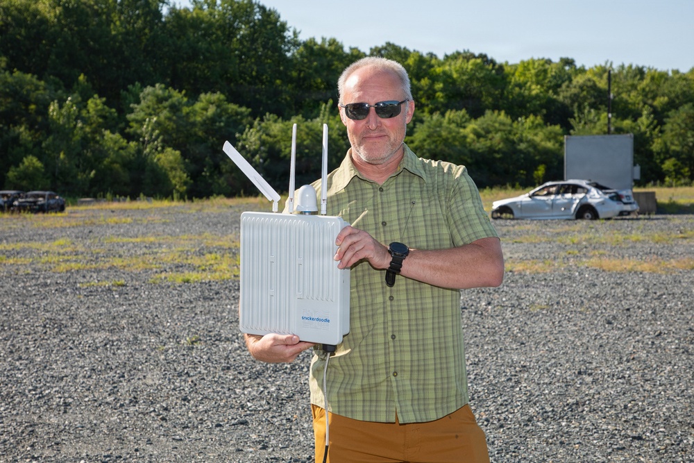 A middle-aged man with a gray beard and sunglasses stands in an open gravel lot holding a white outdoor networking device branded 'snickerdoodle' with multiple antennas. He is wearing a green plaid shirt, mustard-colored pants, and a black watch. The background shows trees, a gravel field, and what appears to be a damaged or wrecked vehicle in the distance.