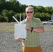 A middle-aged man with a gray beard and sunglasses stands in an open gravel lot holding a white outdoor networking device branded 'snickerdoodle' with multiple antennas. He is wearing a green plaid shirt, mustard-colored pants, and a black watch. The background shows trees, a gravel field, and what appears to be a damaged or wrecked vehicle in the distance.