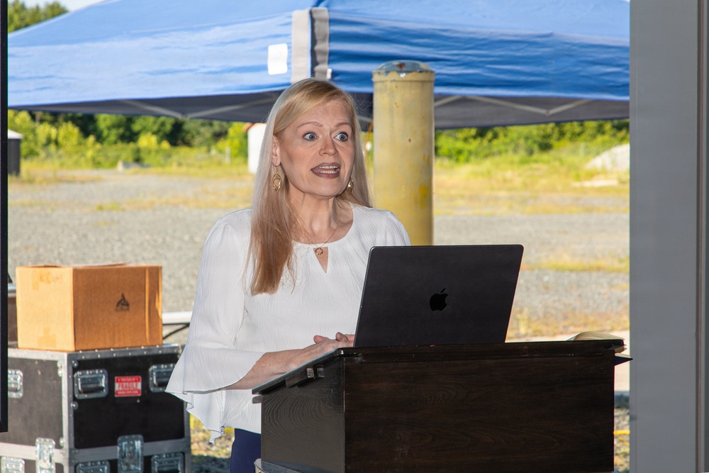 A woman with long blonde hair is speaking at a podium during an outdoor event. She is wearing a white blouse and has a wide-eyed, expressive facial expression while presenting. An Apple MacBook laptop is open on the wooden lectern in front of her.