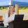 xTechAutonomy Phase II Live Demonstrations A woman with long blonde hair is speaking at a podium during an outdoor event. She is wearing a white blouse and has a wide-eyed, expressive facial expression while presenting. An Apple MacBook laptop is open on the wooden lectern in front of her.