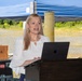 A woman with long blonde hair is speaking at a podium during an outdoor event. She is wearing a white blouse and has a wide-eyed, expressive facial expression while presenting. An Apple MacBook laptop is open on the wooden lectern in front of her.