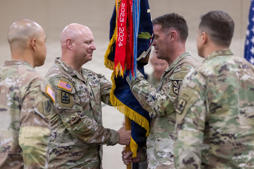 A U.S. Army change of command ceremony is taking place, with two officers exchanging a unit guidon (flag). The ceremony appears to be held indoors in a large facility, with the guidon displaying streamers including a red one marked '2023-2024'. Multiple soldiers in OCP (Operational Camouflage Pattern) uniforms are present as witnesses.