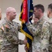 A U.S. Army change of command ceremony is taking place, with two officers exchanging a unit guidon (flag). The ceremony appears to be held indoors in a large facility, with the guidon displaying streamers including a red one marked '2023-2024'. Multiple soldiers in OCP (Operational Camouflage Pattern) uniforms are present as witnesses.