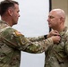 Two U.S. Army soldiers in MultiCam/OCP camouflage uniforms are participating in a military awards or pinning ceremony. One soldier is pinning a medal or decoration onto the chest of the other soldier. The ceremony appears to be taking place indoors against a light-colored backdrop.