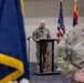 A U.S. Army soldier in OCP (Operational Camouflage Pattern) uniform is speaking at a podium during a formal military ceremony. The setting appears to be an indoor venue with flags including the U.S. flag and what appears to be the Arizona state flag displayed behind the speaker. Other uniformed personnel and a military guidon are visible in the foreground.