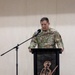 A U.S. Army officer in OCP (Operational Camouflage Pattern) uniform stands at a decorative podium delivering a speech or address. The podium features a laser-cut metal silhouette of a soldier carrying a rifle. A microphone on a stand is positioned to the left of the speaker.