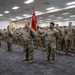 A large formation of U.S. Army soldiers in OCP (Operational Camouflage Pattern) uniforms are assembled indoors in what appears to be a military ceremony or formation event. Two soldiers in the foreground are serving as guidon bearers, holding unit flags including a prominent red guidon marked '487'. The formation is organized in rows within a large indoor facility with fluorescent lighting.