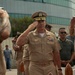 A group of U.S. Navy personnel in Service Khaki (Type B) uniforms are engaged in a formal saluting ceremony outdoors. The central figure, a senior officer with command insignia and multiple service ribbons, is returning a salute from subordinates. The setting appears to be a military installation or official venue with a modern curved glass building in the background.