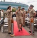 A formal U.S. Navy military ceremony is taking place outdoors, featuring a senior officer walking down a red carpet flanked by decorative torpedo-shaped stanchions while personnel render salutes. The central figure appears to be a high-ranking officer, likely an admiral, distinguished by a gold-brimmed cap and extensive medal ribbons on his uniform. Civilians and uniformed personnel are seated and standing in attendance, with a large modern glass building visible in the background.