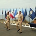 Two U.S. Navy sailors in khaki service uniforms are marching in formation during an outdoor military ceremony. The event is set against a coastal backdrop with a row of state and military flags displayed on staffs. A ceremonial table with Navy insignia, covers (hats), and what appears to be a wooden award or plaque is visible to the right.