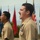 C3F conducts Chief pinning ceremony Two military personnel in tan/khaki dress uniforms are standing at attention and appear to be reciting or singing, possibly during a formal ceremony. Multiple flags are visible in the background, including the American flag, POW/MIA flag, and various military branch flags. The foreground subject displays multiple service ribbons and medals on his uniform.
