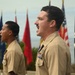 Two military personnel in tan/khaki dress uniforms are standing at attention and appear to be reciting or singing, possibly during a formal ceremony. Multiple flags are visible in the background, including the American flag, POW/MIA flag, and various military branch flags. The foreground subject displays multiple service ribbons and medals on his uniform.