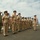C3F conducts Chief pinning ceremony A formal U.S. Navy military ceremony is taking place outdoors on a paved surface, likely a parade ground or rooftop area with an ocean or large body of water visible in the background. A formation of Navy personnel in tan/khaki service uniforms (Service Uniform Bravo) stand at attention, with one individual in NWU (Navy Working Uniform) camouflage at the far left. Multiple military flags and guidons are displayed on both sides of the formation.