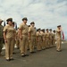 A formal U.S. Navy military ceremony is taking place outdoors on a paved surface, likely a parade ground or rooftop area with an ocean or large body of water visible in the background. A formation of Navy personnel in tan/khaki service uniforms (Service Uniform Bravo) stand at attention, with one individual in NWU (Navy Working Uniform) camouflage at the far left. Multiple military flags and guidons are displayed on both sides of the formation.