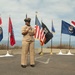 A U.S. Navy enlisted service member in khaki service uniform stands at a microphone addressing an audience at an outdoor military ceremony. The setting appears to be a coastal overlook or memorial area with multiple state flags, the U.S. national flag, and a POW/MIA flag displayed on poles behind the speaker. The ocean is visible in the background under a partly cloudy sky.